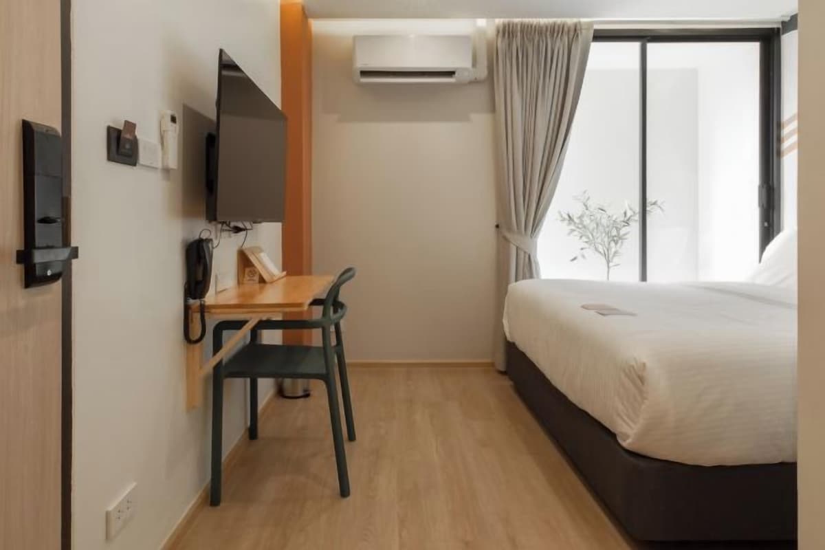 A hotel room with a bed, a desk with a chair, a TV, an air conditioner, and a window with curtains.