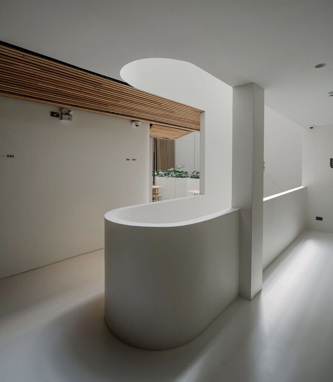 Un área de recepción moderna y minimalista con un mostrador blanco curvo, techo de listones de madera y un muro espejado que refleja un baño con lavamanos y plantas.