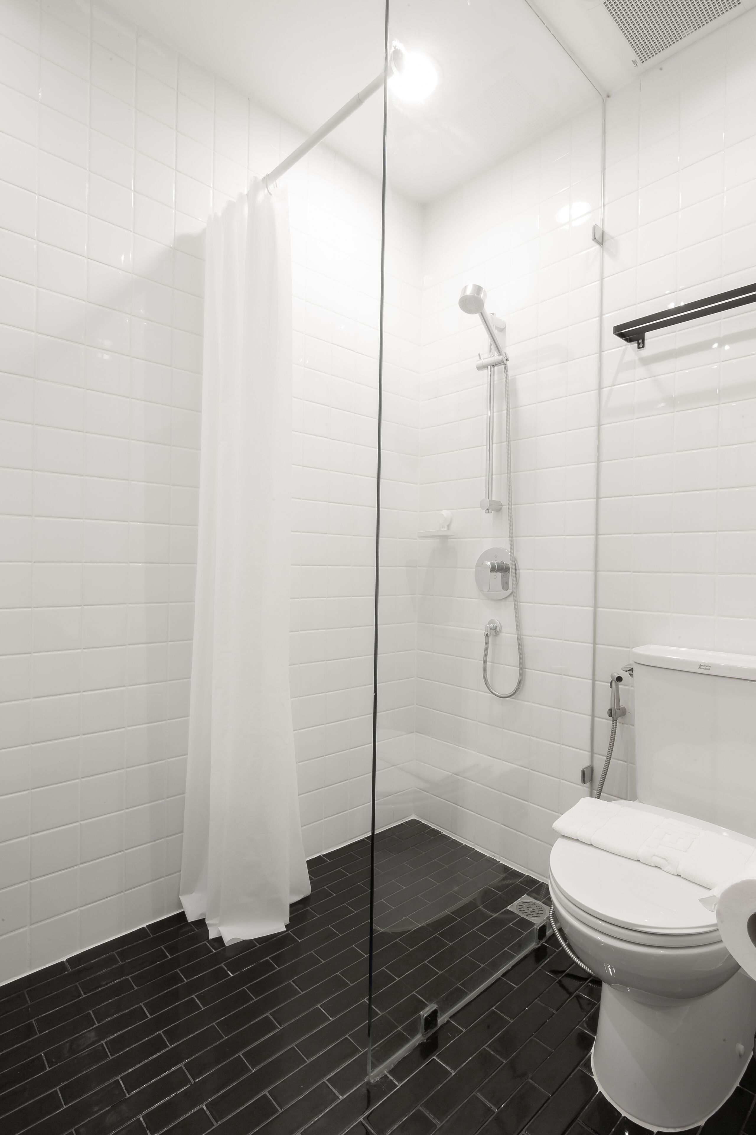 A modern bathroom with a glass-enclosed shower, white tiled walls, a black tiled floor, and a white toilet.