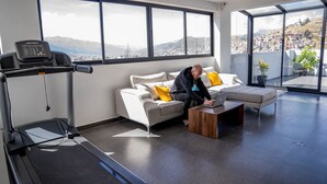 A modern living room with a treadmill, a sofa with yellow pillows, a coffee table, and a city view through large windows. - Spacious 4 Bedroom Full Apartment, a few Minutes From the Main Square (Cusco)