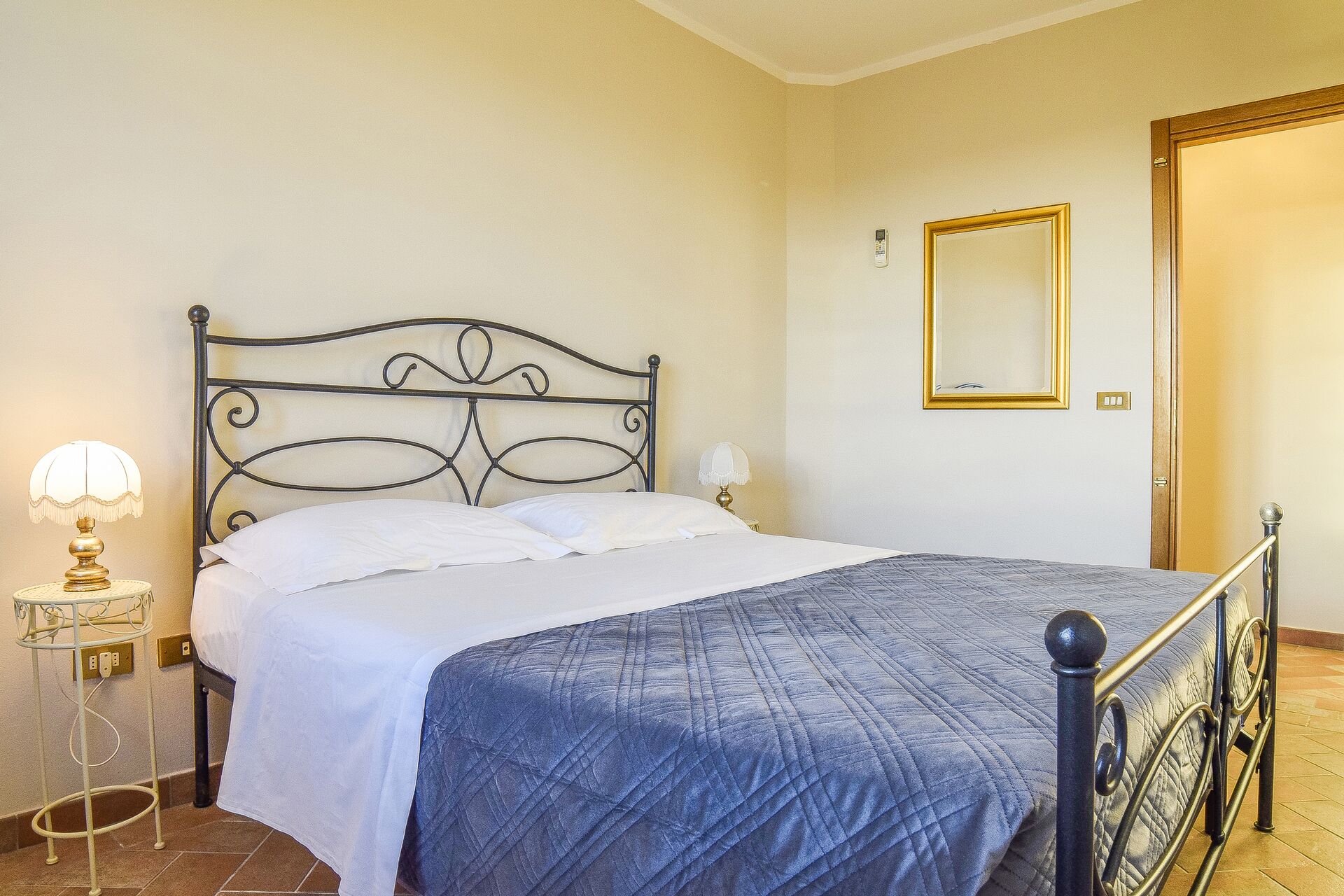 Una cama bien hecha con un edredón azul, una mesita de noche con lámpara y un espejo en la pared.