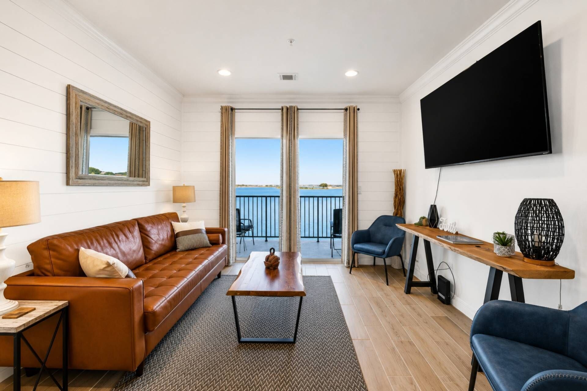A living room with a brown leather sofa, a wooden coffee table, and a blue armchair. A flat-screen TV is mounted on the wall, and there is a balcony with a view of the water.