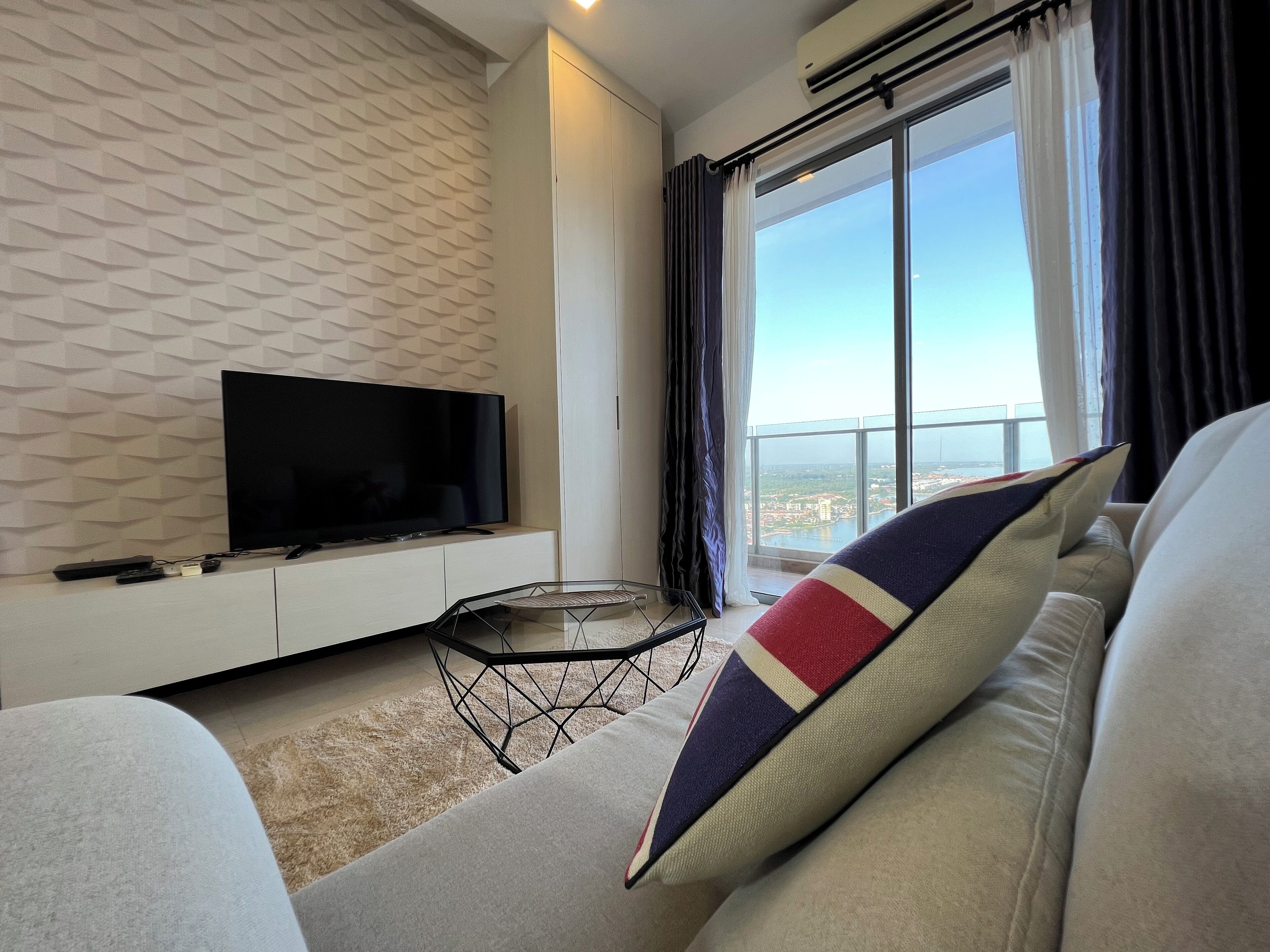 A modern living room with a flat-screen TV on a white cabinet, a glass coffee table, and a sofa with a colorful pillow.