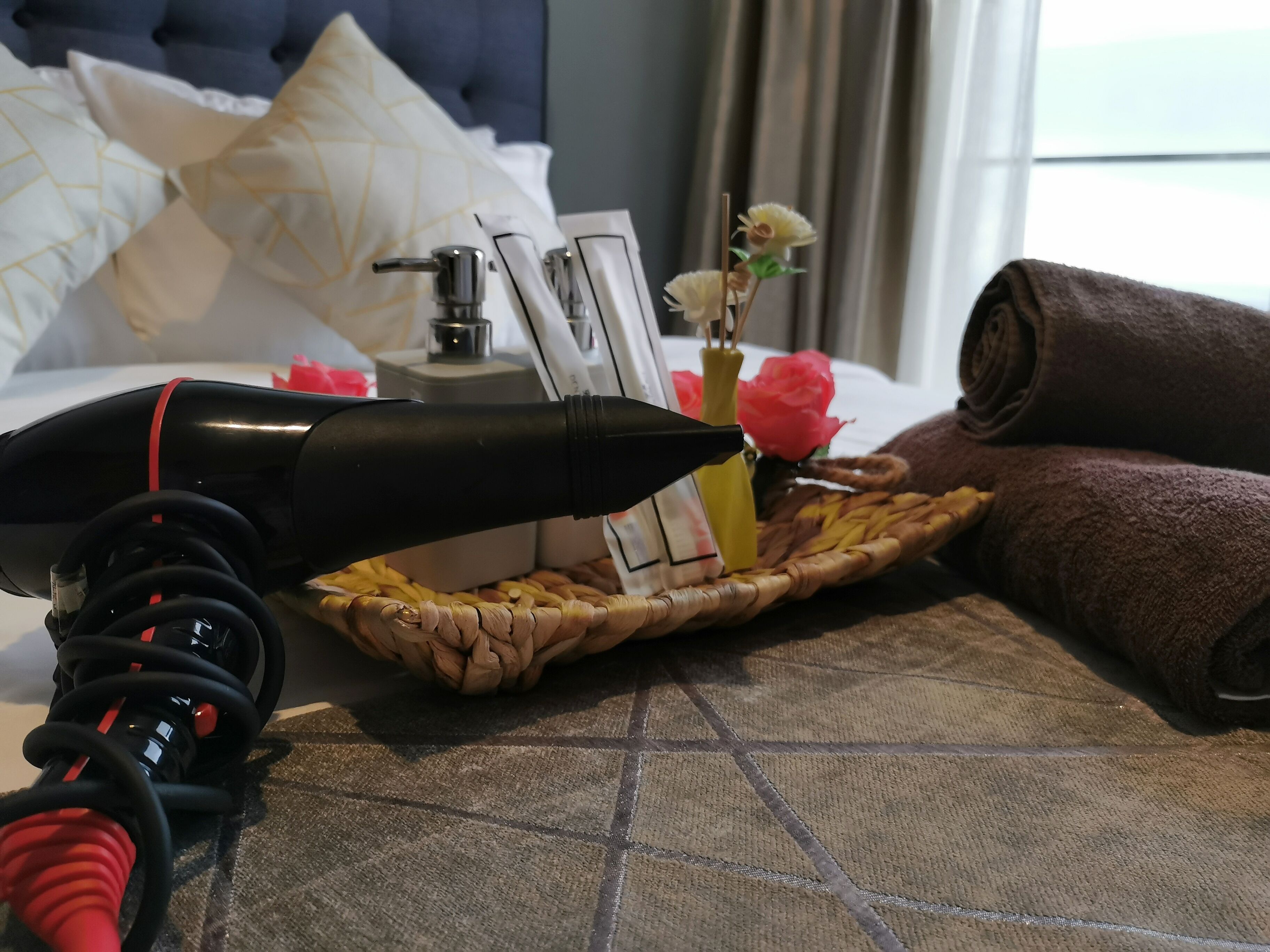 Habitación de hotel ordenada con cama, almohadas, mesita de noche con lámpara, toalla enrollada y una cesta decorativa con flores.