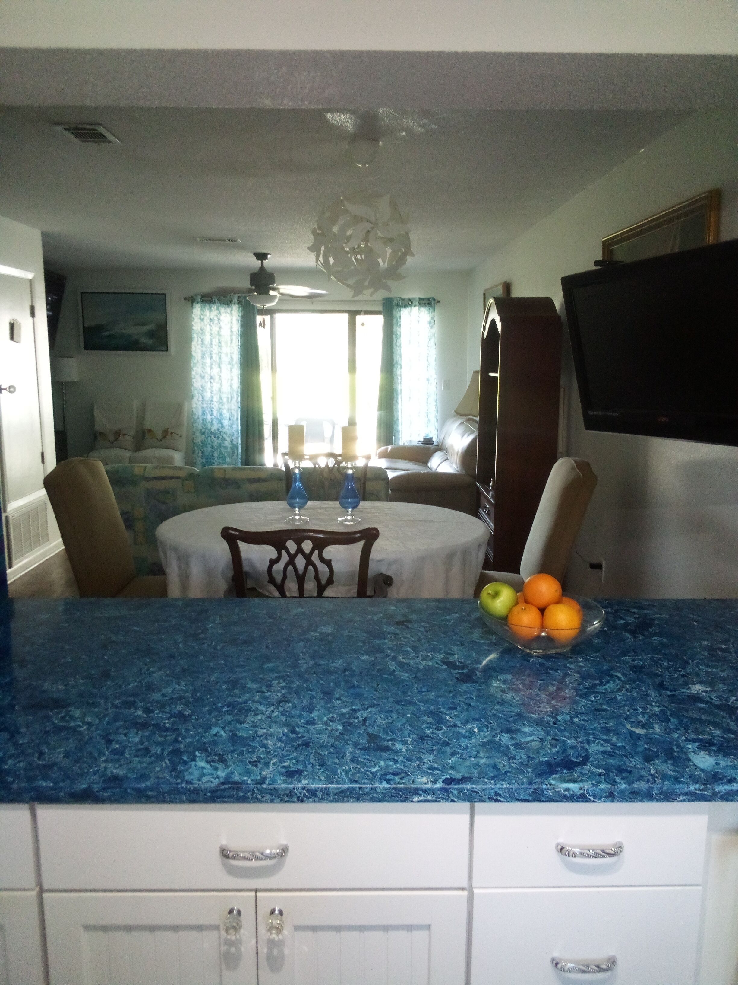 A kitchen with a blue countertop, a dining table with chairs, a TV mounted on the wall, and a fruit bowl on the counter.