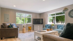 Un salon avec un canapé marron, une table basse blanche et un fauteuil bleu. On y trouve un téléviseur posé sur un meuble blanc, ainsi qu’une fenêtre avec des stores.