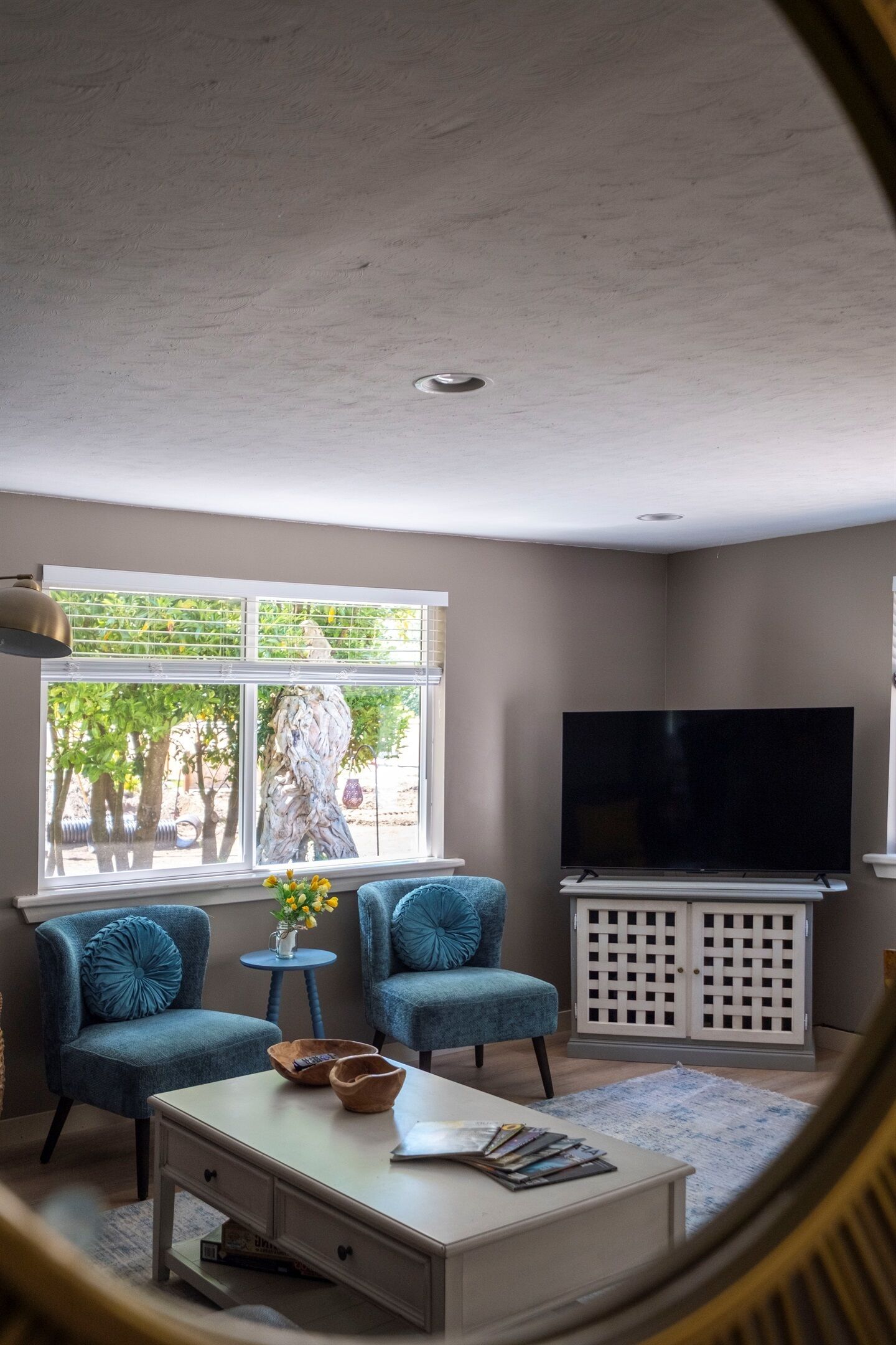 Een woonkamer met een grote televisie, twee blauwe fauteuils, een witte salontafel en een raam met uitzicht op bomen.