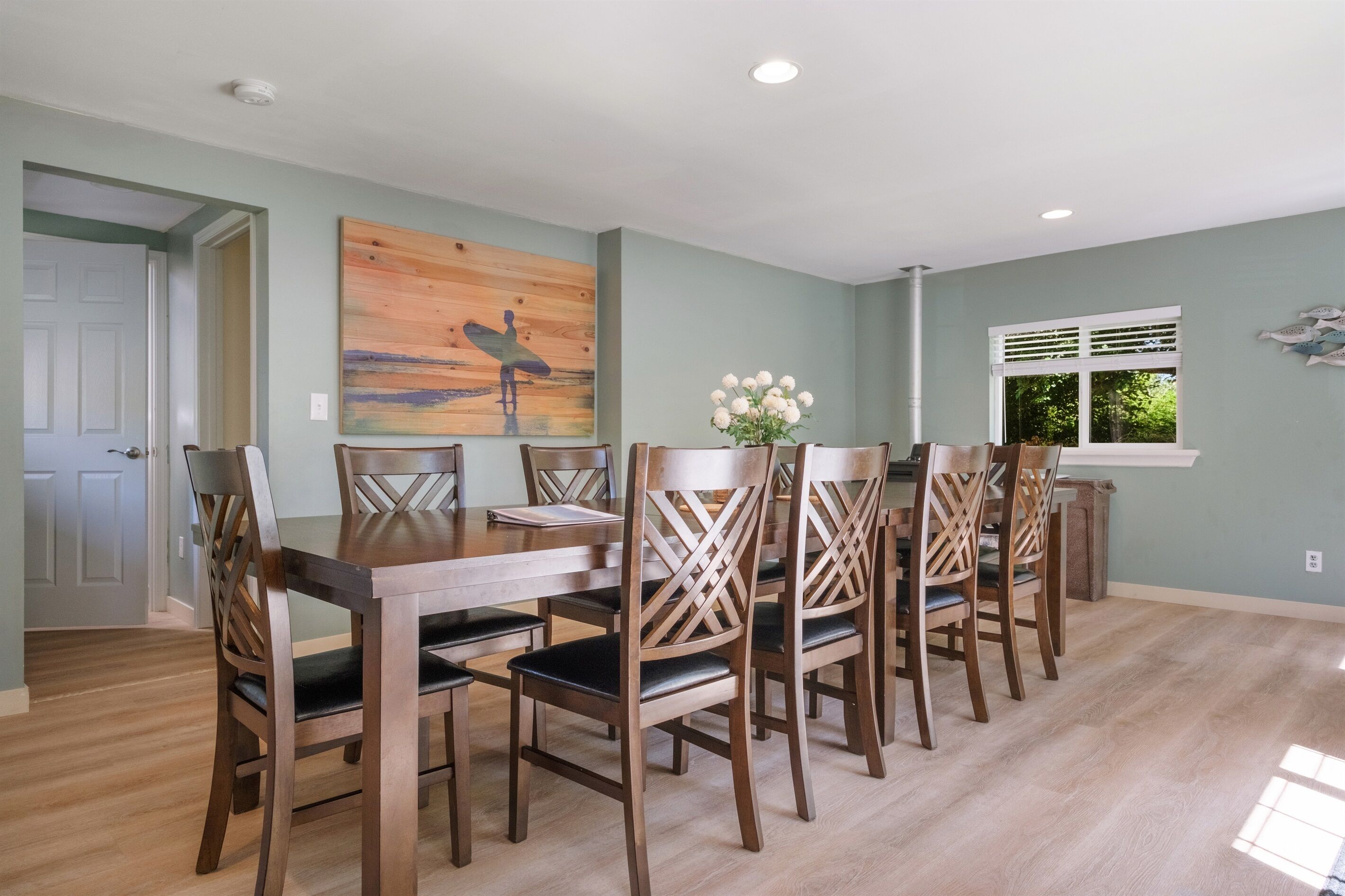 A dining area with a wooden table and chairs, a painting on the wall, and a window with blinds.
