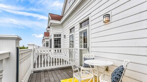 Una terraza de madera con barandas blancas, un tapete amarillo estampado y sillas de metal blanco con cojines decorados.