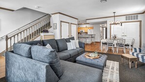 Un salón amplio con un sofá seccional gris, un banco de madera y una zona de comedor con una mesa y sillas blancas.