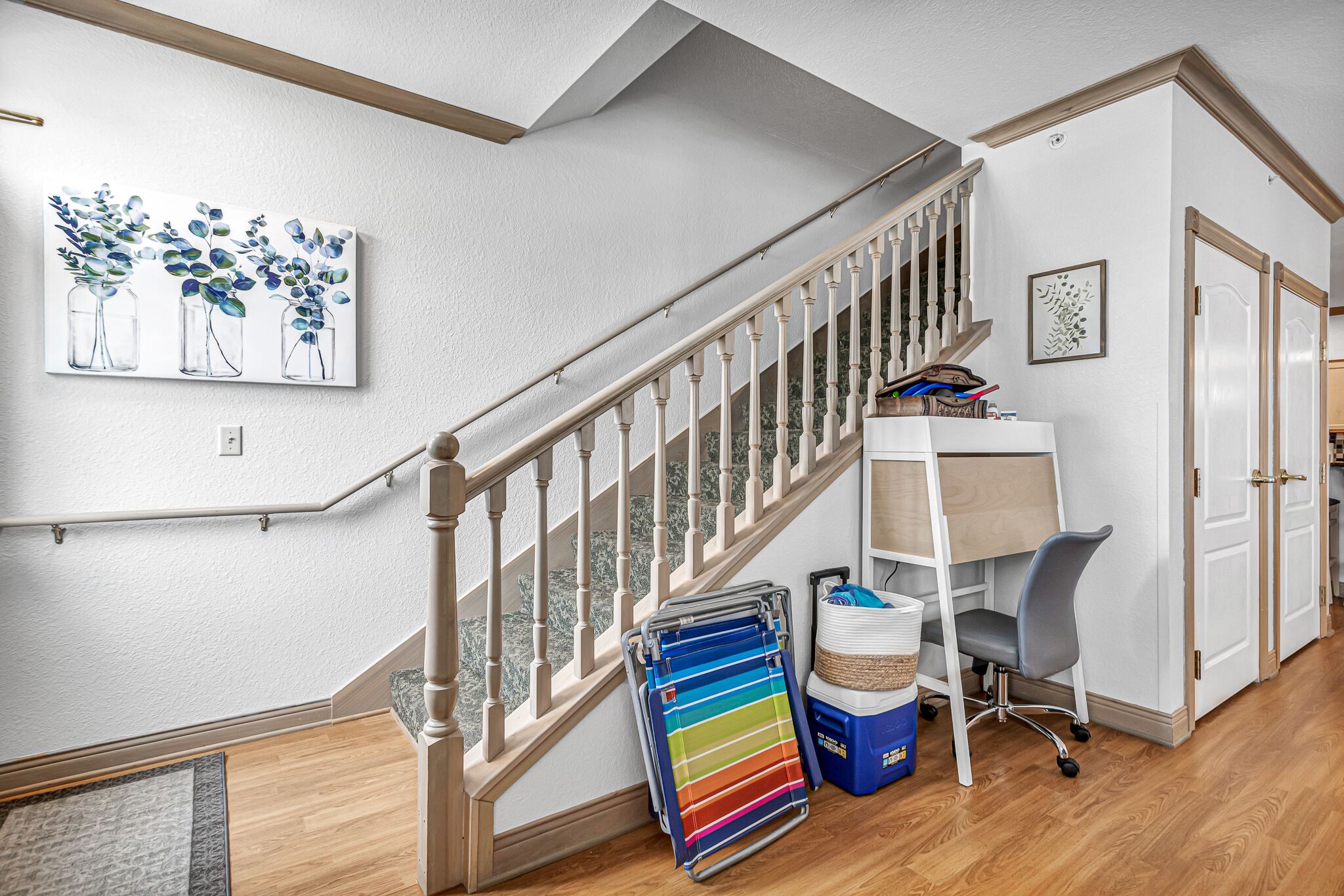 A staircase with a wooden handrail, a painting of vases with flowers on the wall, a desk with a chair, and a colorful towel rack.