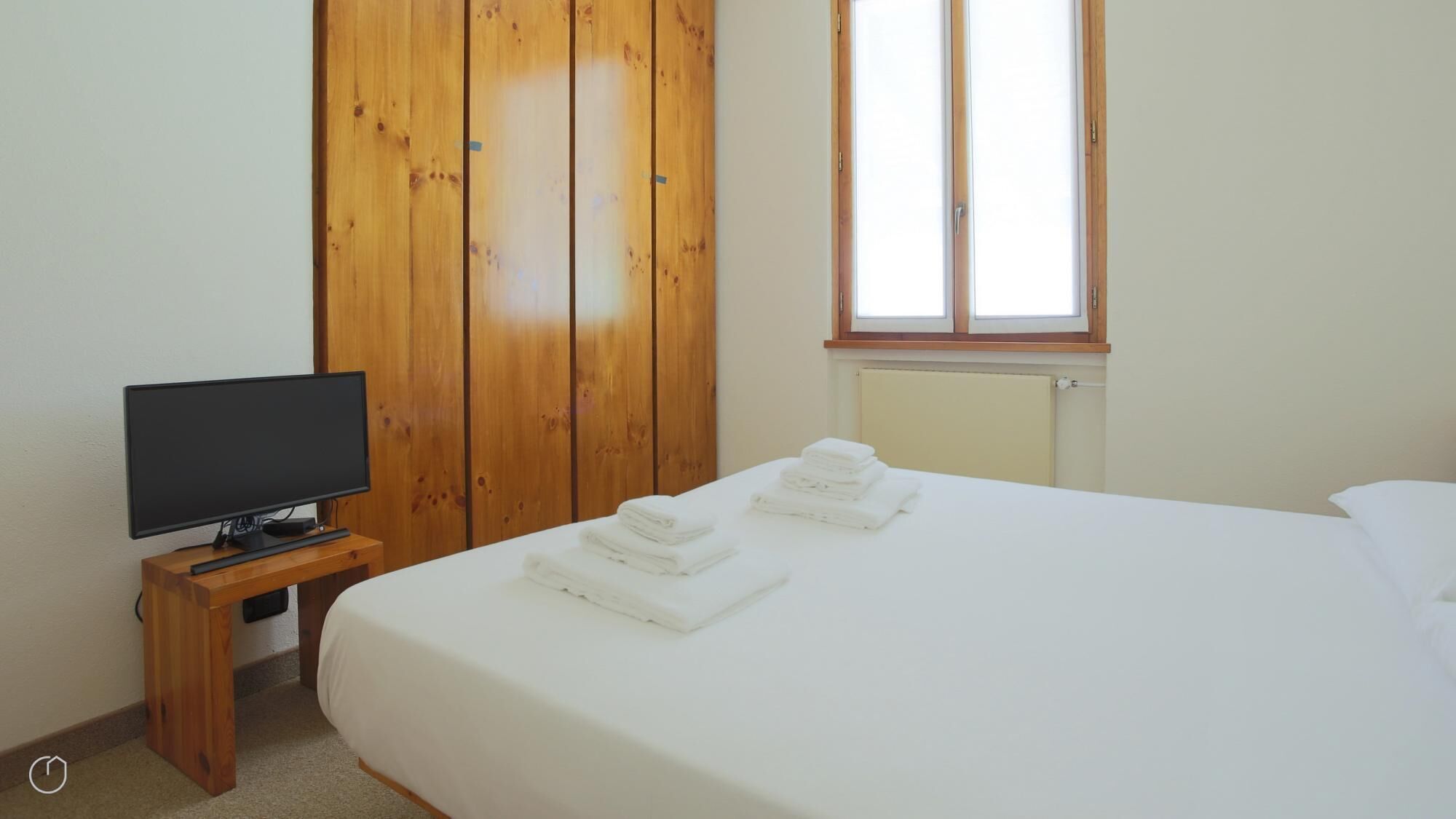 A bedroom with a bed, a television on a wooden stand, a window, and a radiator.