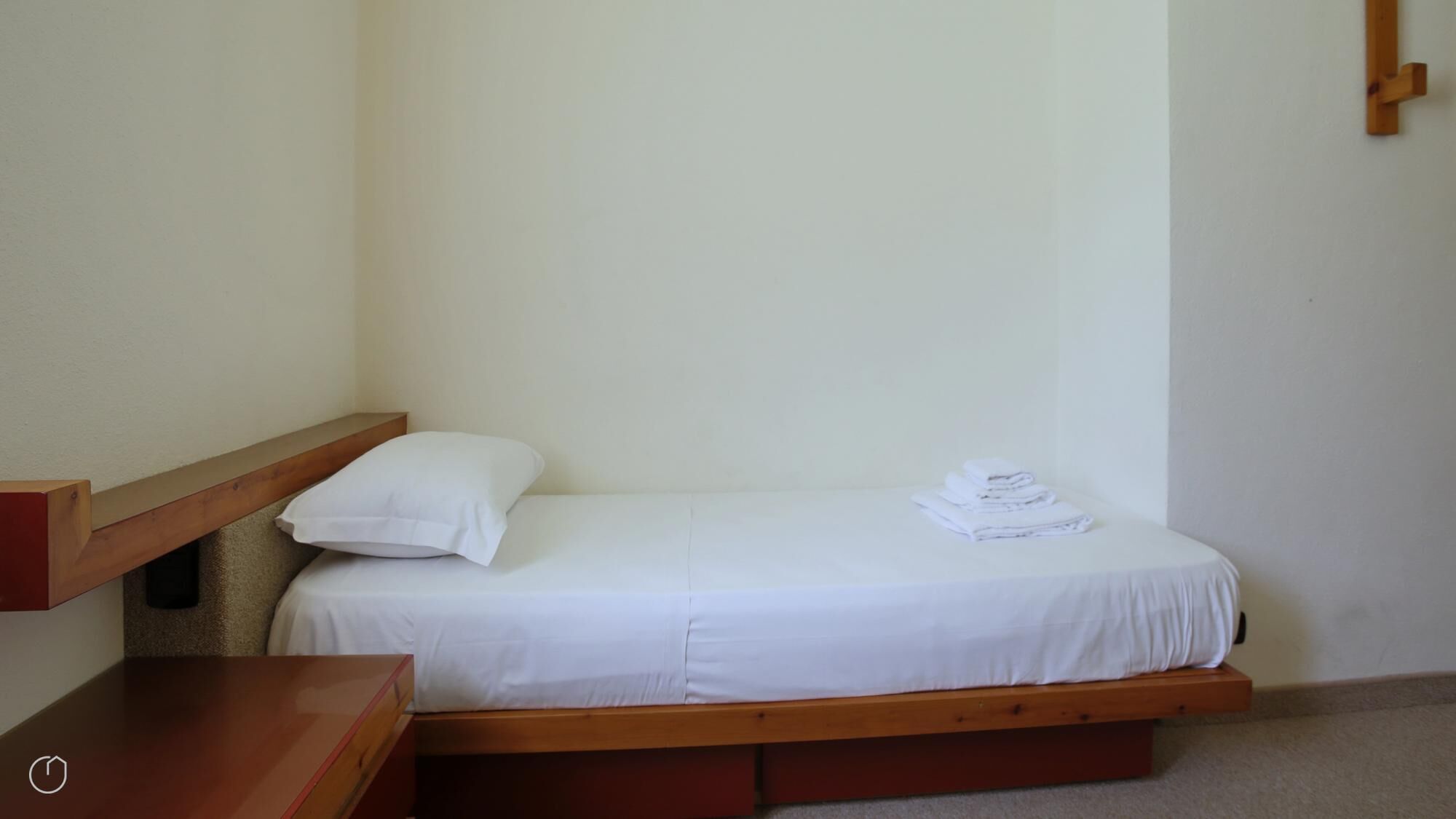 A small, single-bed room with a wooden frame, white bedding, and a small table.