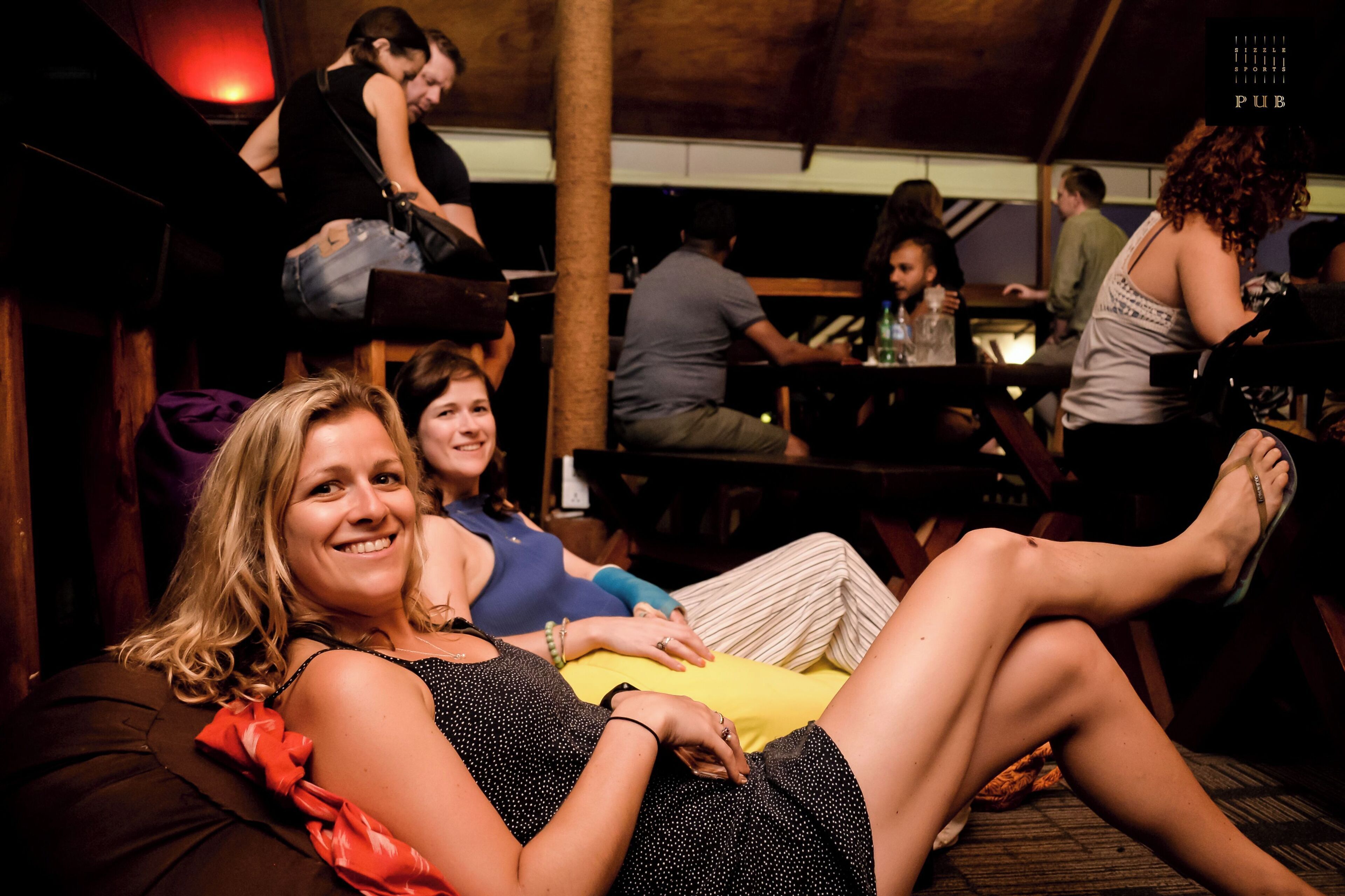 A group of people sitting and standing in a dimly lit indoor setting with wooden furniture and tables.