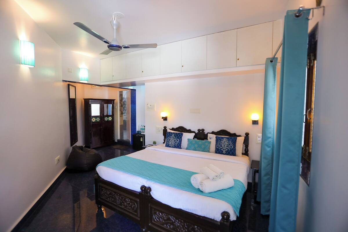Quarto de hotel com cama, ventilador de teto, mesa de cabeceira, luminária e porta do banheiro.