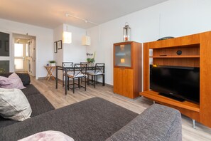 A modern living room with a sofa, TV, wooden entertainment unit, dining area, and a door leading to another room.