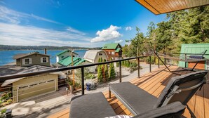 A balcony with wooden flooring, a hammock, and a view of a lake and houses. - Beach Access, Chef’s Kitchen, Rain Shower, Peloton (Camano)