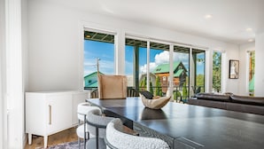A modern dining area with a large table, cushioned chairs, and a view of a green-roofed house through large windows. - Beach Access, Chef’s Kitchen, Rain Shower, Peloton (Camano)