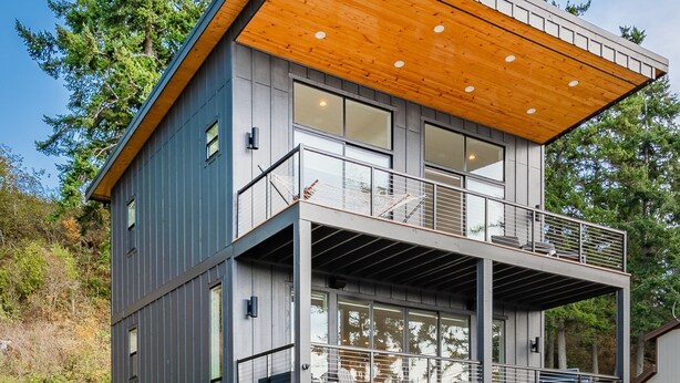 A modern two-story house with a wooden deck, large windows, and a balcony. - Beach Access, Chef’s Kitchen, Rain Shower, Peloton (Camano)