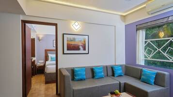 A modern hotel room with a grey sofa, a framed picture on the wall, a ceiling fan, and a window with a view of greenery.
