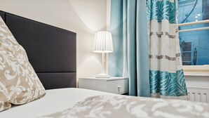 A hotel room with a bed, a nightstand with a lamp, a window with curtains, and a radiator. - Florian's apartments (Vienna)