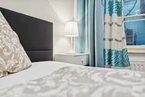 A hotel room with a bed, a nightstand with a lamp, a window with curtains, and a radiator.
