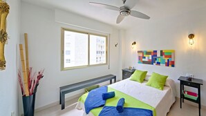 A modern bedroom with a bed, a ceiling fan, a window, a bench, a bedside table, and a colorful abstract painting.