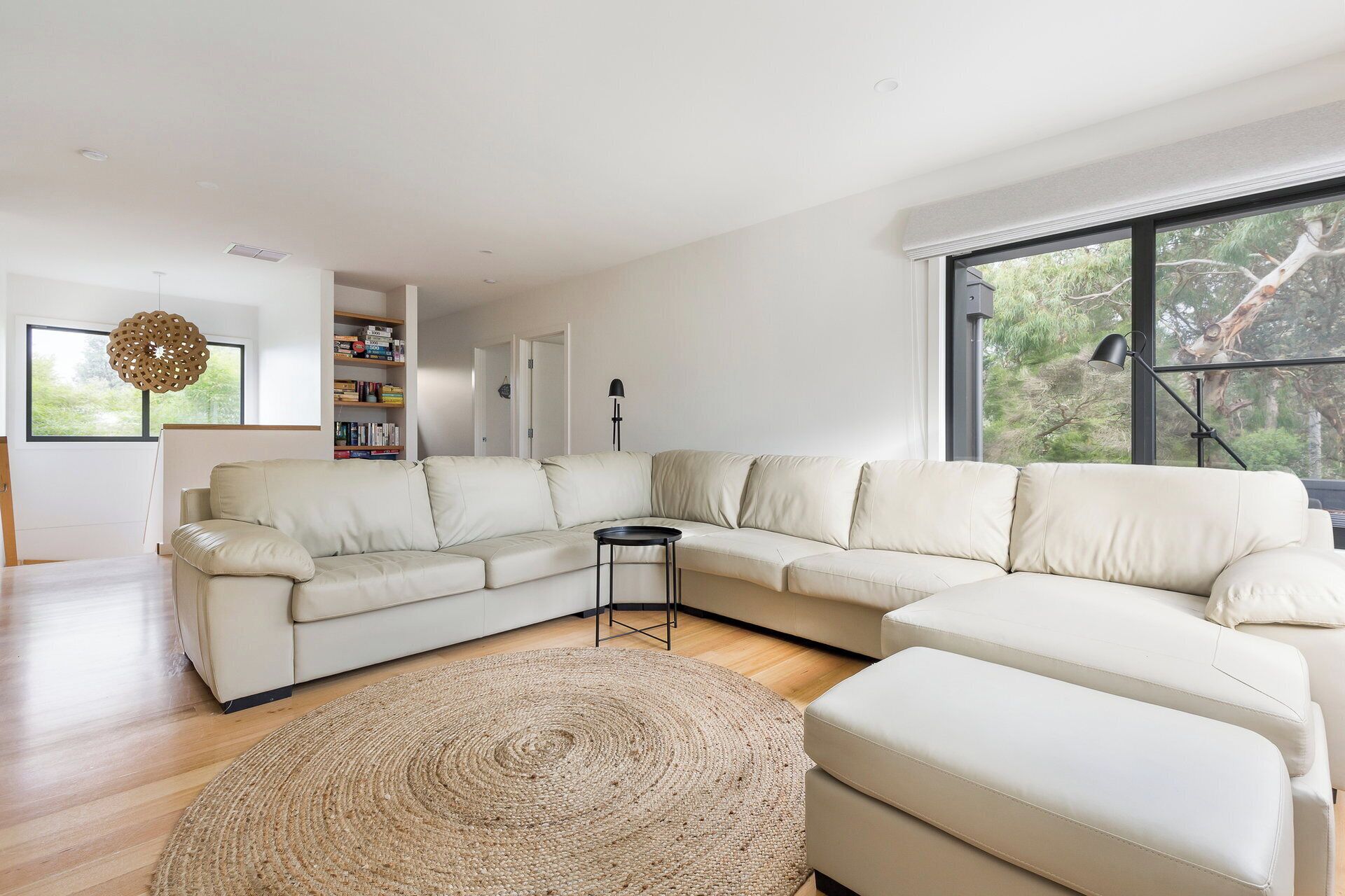Een moderne woonkamer met een hoekbank, een rond geweven vloerkleed, een boekenkast en een groot raam met uitzicht op bomen.