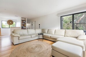Ein modernes Wohnzimmer mit einer Eckcouch, einem runden Teppich, einem Bücherregal und einem großen Fenster mit Blick auf Bäume.