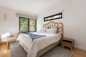 Ein Schlafzimmer mit einem großen Bett, einem Sessel, einem kleinen Tisch, einem Nachttisch und einem Fenster mit Blick auf Bäume.