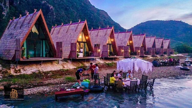 A row of wooden cabins with triangular roofs by a river, some with windows open, and a dining area with tables and chairs.