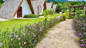 A row of triangular-roofed cottages with a dirt path leading through a garden with purple flowers.