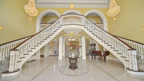 Una gran escalera con barandales blancos y suelo pulido que conduce a un área central con un piano y un arreglo floral.