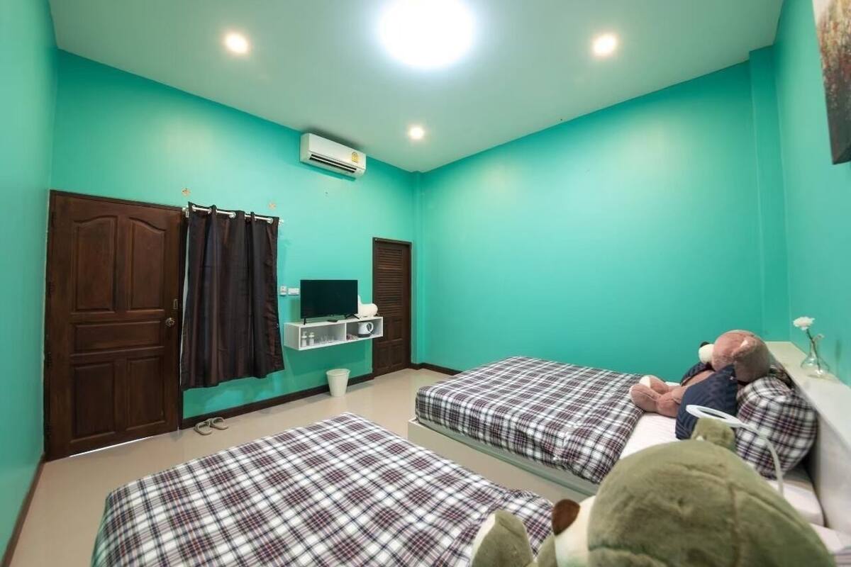 Une pièce aux murs de couleur turquoise, avec deux lits recouverts de draps à carreaux, une porte en bois, une petite table avec une chaise, un téléviseur et un appareil de climatisation.