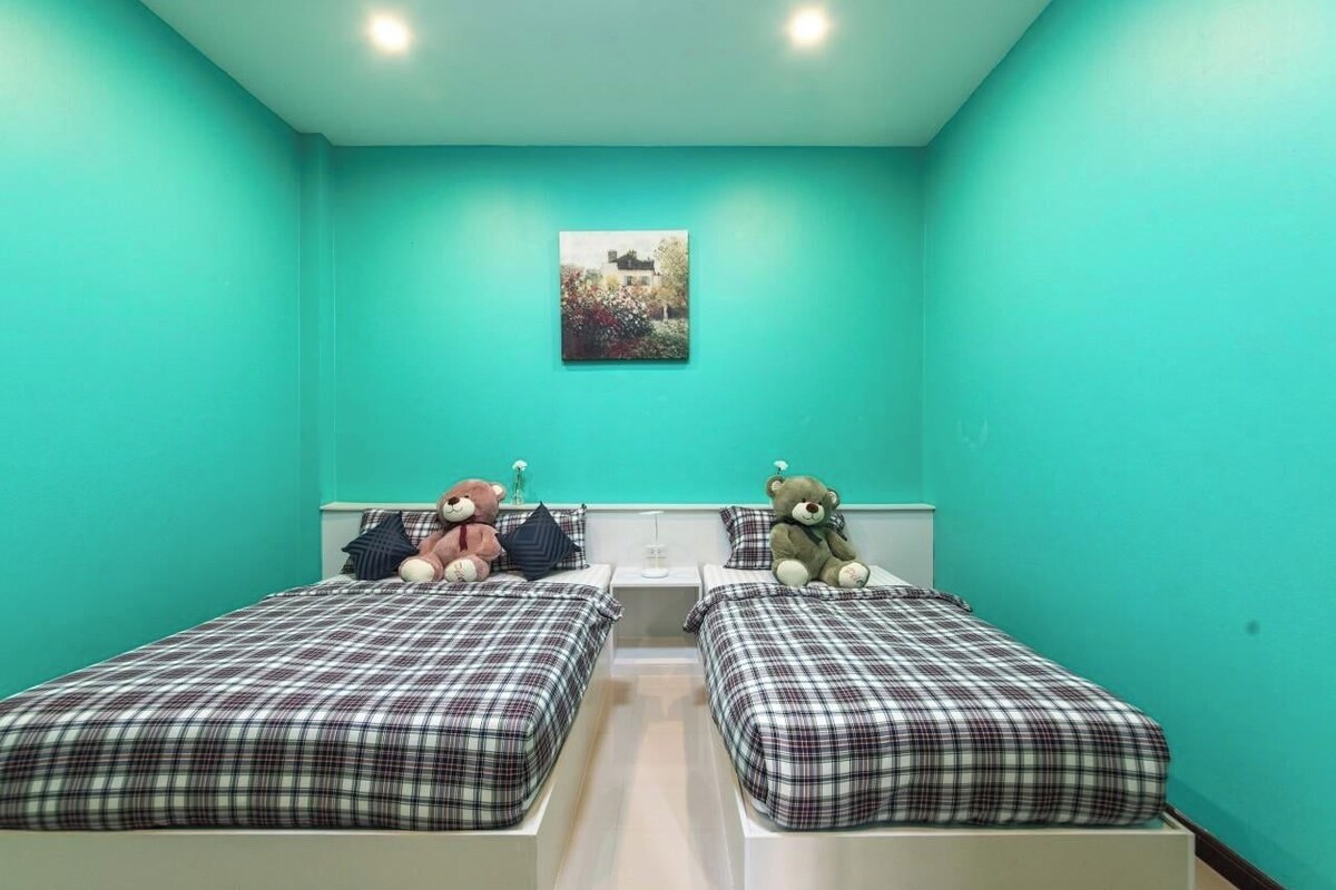 Ein Zimmer mit zwei Betten, jeweils mit kariertem Überwurf, einem kleinen Tisch in der Mitte und einem an der Wand hängenden Bild.