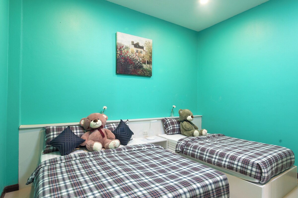 A room with teal walls, two beds with plaid bedding, a white nightstand, and a teddy bear on each bed.