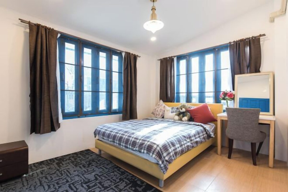 A bedroom with a bed, a desk, and a chair. There are windows with curtains.