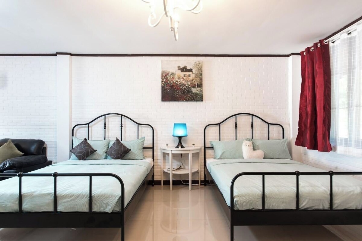 A room with two beds, a small round table with a lamp, a black leather sofa, and a window with red curtains.