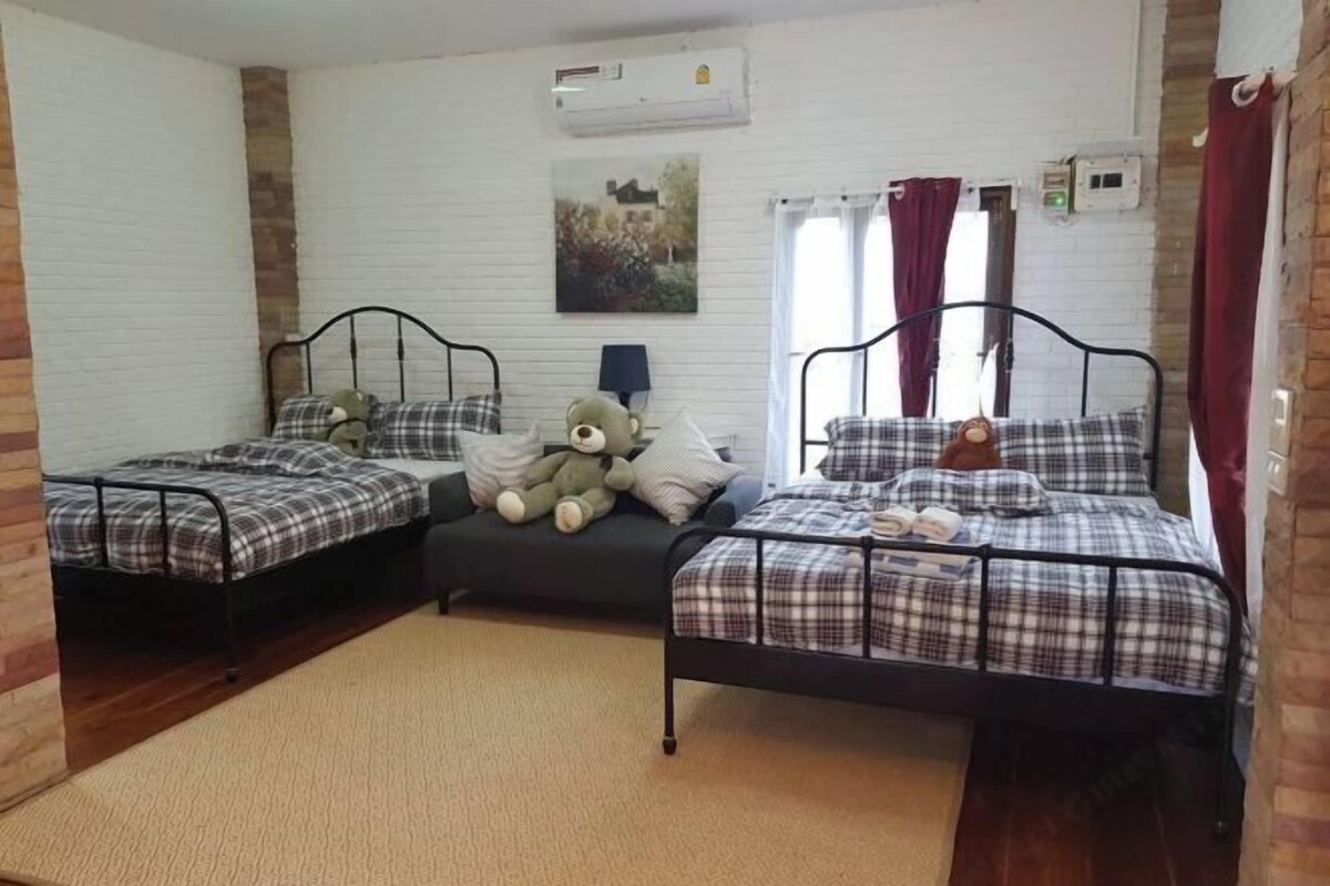 Una habitación con dos camas, cada una con estructura metálica y ropa de cama escocesa. Hay un oso de peluche grande en una cama, y una lámpara en la otra. Un cuadro cuelga de la pared, y hay una ventana con cortinas.