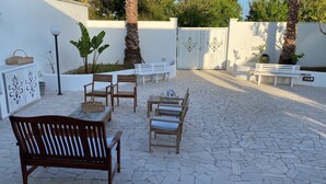 Un patio exterior con suelo de piedra, bancos de madera y una pared blanca con paneles decorativos.