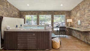 A stone-walled room with a hot tub, wooden flooring, a table with chairs, and a view of greenery outside.