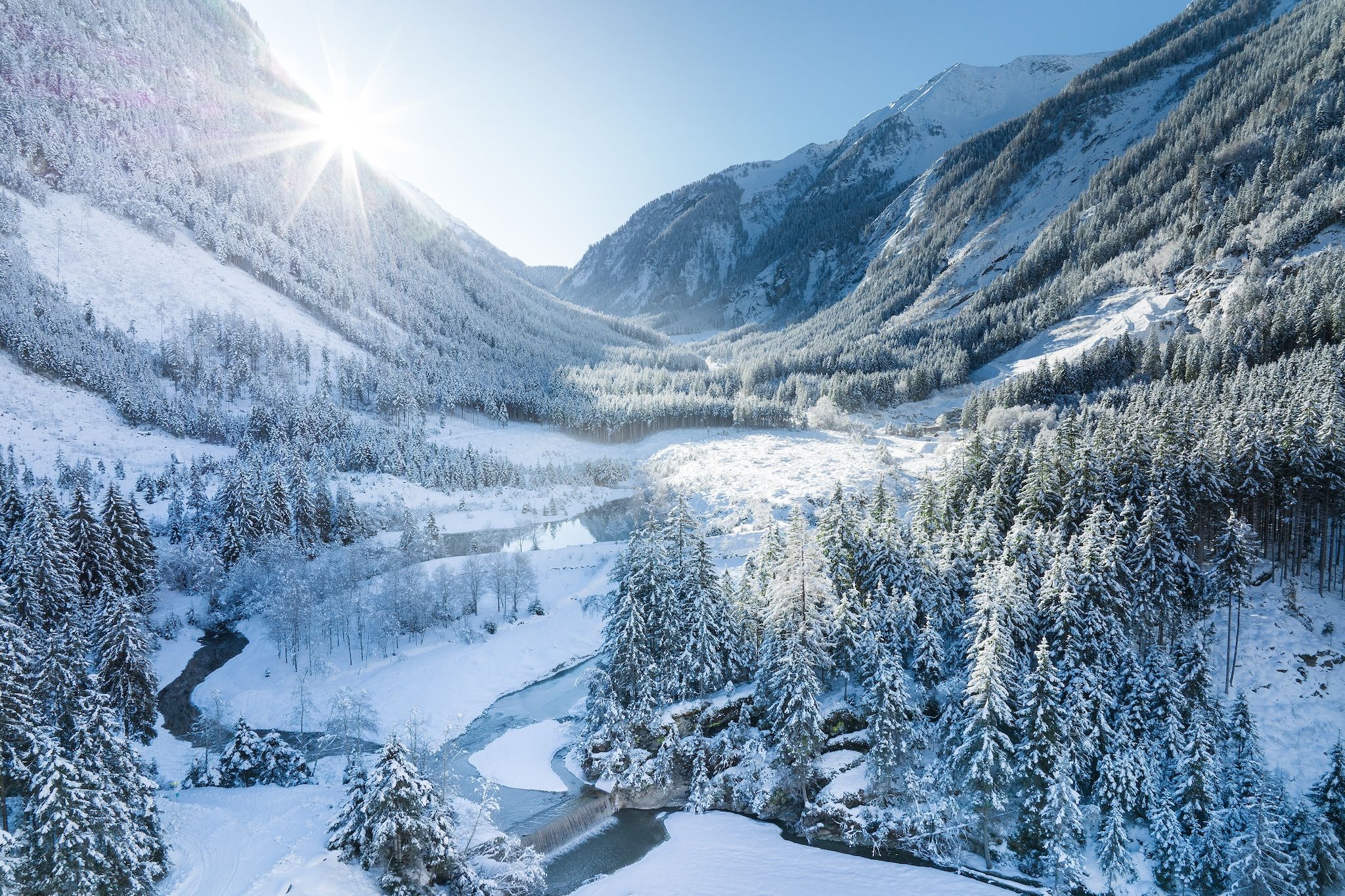 A snow-covered valley with a river, surrounded by dense forests and mountains.