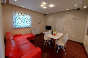 A modern living room with a red sofa, a dining table with chairs, a flat-screen TV mounted on the wall, and a window with a floral pattern.