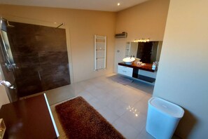 Un baño moderno con cabina de ducha de vidrio, tocador blanco con un espejo grande y piso de baldosas color beige.