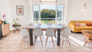 A bright dining area with a large table set for four, a sofa, and a view of greenery outside. - Grande Villa à 400m de la Plage, Idéale Famille par Groom (Ploemeur)