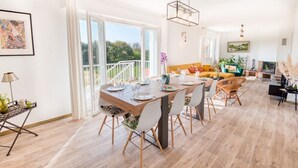 A spacious dining area with a wooden table and chairs, a balcony with a railing, and a living area with a sofa and a TV. - Grande Villa à 400m de la Plage, Idéale Famille par Groom (Ploemeur)