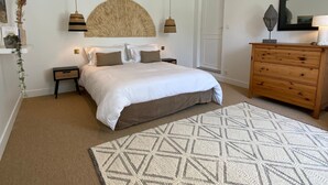 A bedroom with a large bed, bedside tables, a wooden dresser, a mirror, and a patterned rug. - Apartment 'Prat Bras - T1 Cocooning' with Shared Garden and Wi-Fi (Clohars-Carnoët)