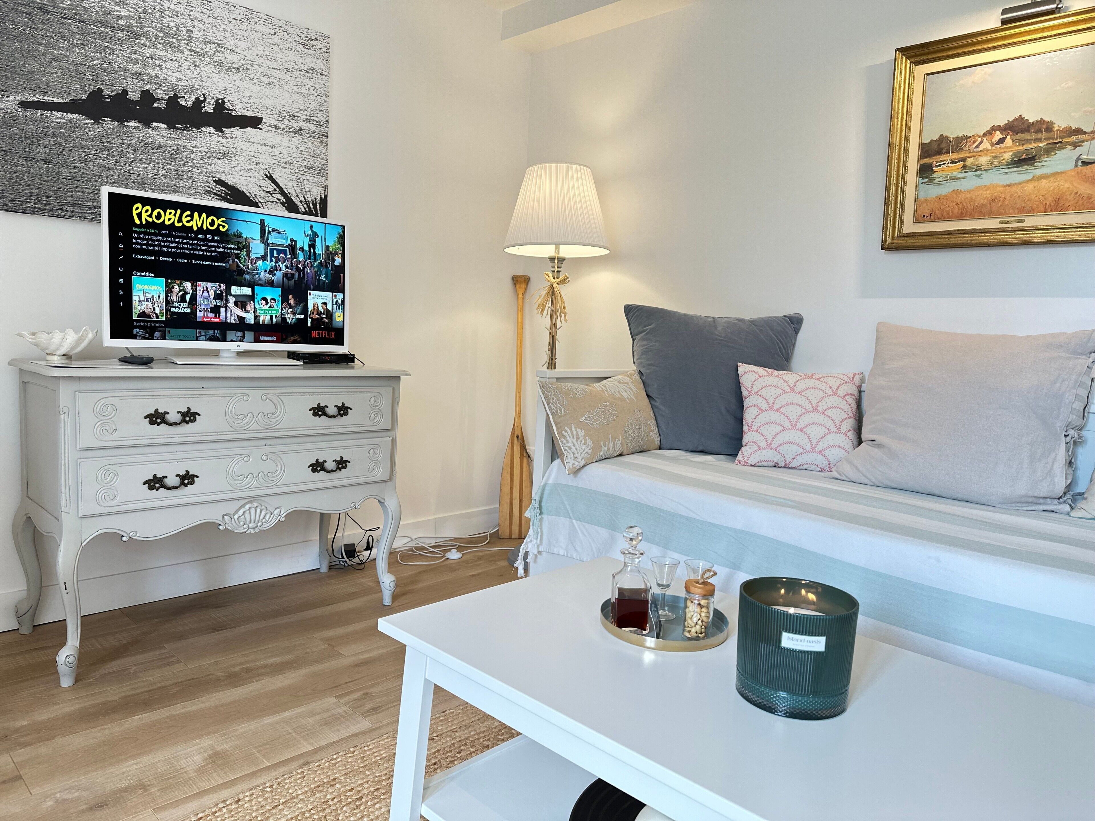 Un salon avec un téléviseur affichant l’interface de Netflix, une commode blanche, un canapé avec des coussins, une lampe et un tableau accroché au mur.