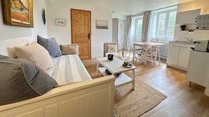 A cozy living room with a sofa, a dining table, and a kitchen area with cabinets and a window. - Apartment 'Prat Bras - T1 Cocooning' with Shared Garden and Wi-Fi (Clohars-Carnoët)