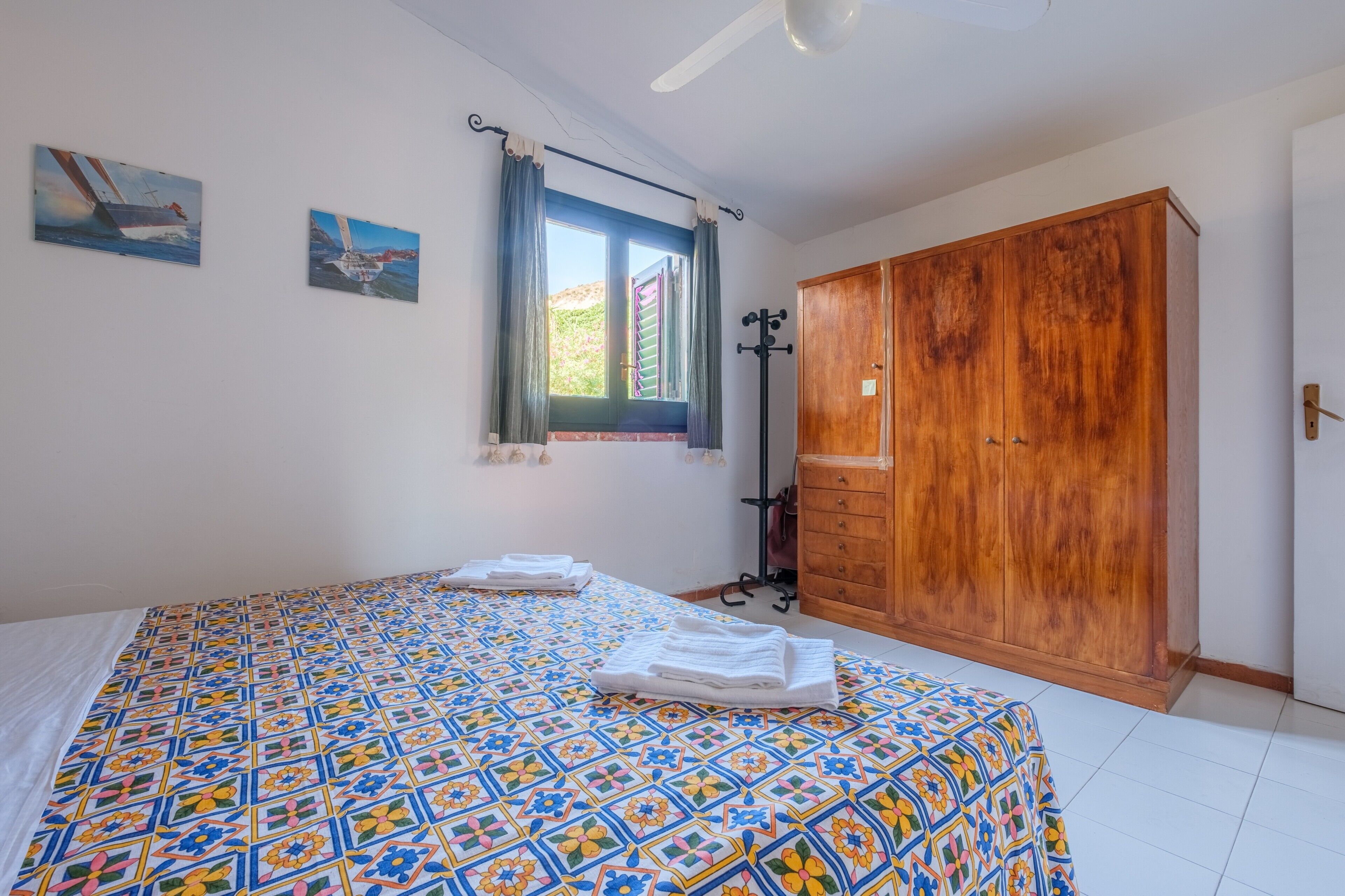 Una camera da letto con un letto, un armadio, una finestra con tende e due quadri appesi al muro.